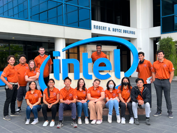 A group of happy students wearing orange polo shirts standing around the Intel sign at the Robert N. Noyce Building (Intel Headquarters in Santa Clara, California)